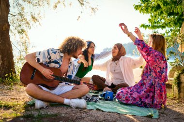 Bir grup arkadaş plajda toplanıp müzik, kahkaha ve birlikteliğin sıcaklığıyla dolu neşeli anları paylaşıyorlar. Gençler sahilde eğleniyor.