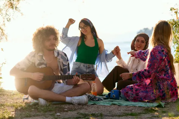 Bir grup arkadaş plajda toplanıp müzik, kahkaha ve birlikteliğin sıcaklığıyla dolu neşeli anları paylaşıyorlar. Gençler sahilde eğleniyor.