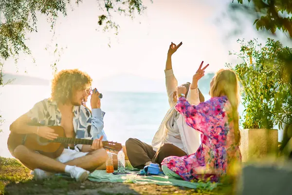 Bir grup arkadaş plajda toplanıp müzik, kahkaha ve birlikteliğin sıcaklığıyla dolu neşeli anları paylaşıyorlar. Gençler sahilde eğleniyor.