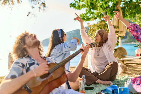 Bir grup arkadaş plajda toplanıp müzik, kahkaha ve birlikteliğin sıcaklığıyla dolu neşeli anları paylaşıyorlar. Gençler sahilde eğleniyor.