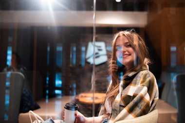 Modern bir kafe ortamında kahve içerken cam duvarın arkasında gülümseyen kareli ceket giyen şık genç bir kadın..