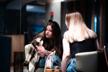 Modern bir kafede, rahat bir atmosferi olan iki genç kadının birbirleriyle etkileşim kurup, akıllı bir telefona baktıkları görüntüler..