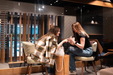 Modern bir kafede, rahat bir atmosferi olan iki genç kadının birbirleriyle etkileşim kurup, akıllı bir telefona baktıkları görüntüler..