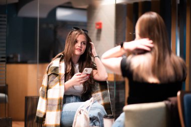 Modern bir kafede, rahat bir atmosferi olan iki genç kadının birbirleriyle etkileşim kurup, akıllı bir telefona baktıkları görüntüler..