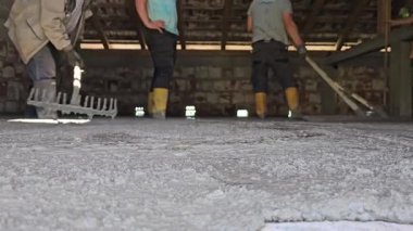 Hakiki hayat insanları sarı çizmeli işçiler inşaat alanındaki tırmıkları kullanarak yeni dökülmüş betonu dümdüz ediyorlar. Düşük açıyla ateş edilmiş, takım çalışmasını ve el işçiliğini gösteriyor