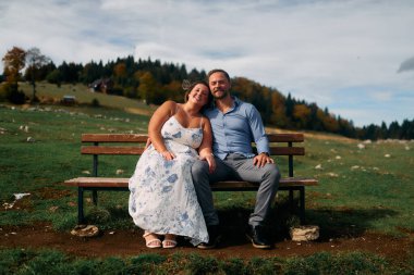 Romantik çift, manzaralı bir dağ çayırında bankta oturuyor, gülümsüyor ve birlikte huzurlu doğanın tadını çıkarıyorlar..