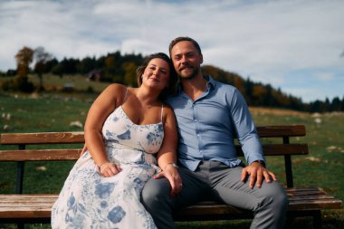 Romantik çift, manzaralı bir dağ çayırında bankta oturuyor, gülümsüyor ve birlikte huzurlu doğanın tadını çıkarıyorlar..