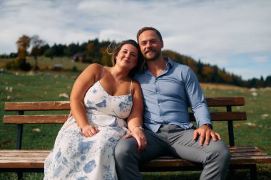 Romantik çift, manzaralı bir dağ çayırında bankta oturuyor, gülümsüyor ve birlikte huzurlu doğanın tadını çıkarıyorlar..