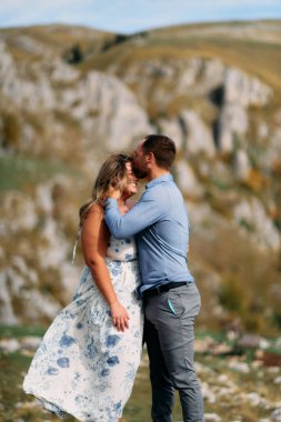 Rüzgarlı bir günde, dağlarda romantik bir anda gülümseyen kadını alnından öpen bir adam..