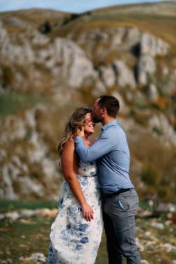 Rüzgarlı bir günde, dağlarda romantik bir anda gülümseyen kadını alnından öpen bir adam..