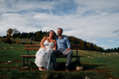 Romantik çift, manzaralı bir dağ çayırında bankta oturuyor, gülümsüyor ve birlikte huzurlu doğanın tadını çıkarıyorlar..