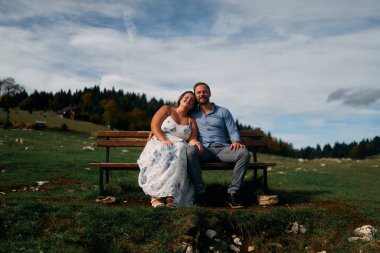 Romantik çift, manzaralı bir dağ çayırında bankta oturuyor, gülümsüyor ve birlikte huzurlu doğanın tadını çıkarıyorlar..