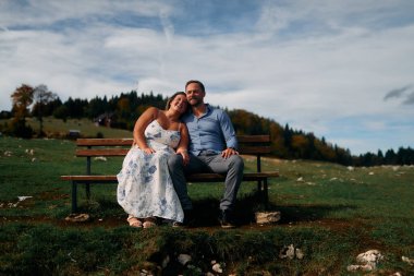 Romantik çift, manzaralı bir dağ çayırında bankta oturuyor, gülümsüyor ve birlikte huzurlu doğanın tadını çıkarıyorlar..