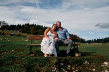 Romantik çift, manzaralı bir dağ çayırında bankta oturuyor, gülümsüyor ve birlikte huzurlu doğanın tadını çıkarıyorlar..