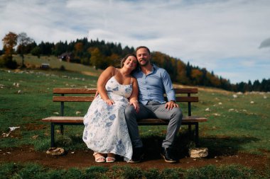 Romantik çift, manzaralı bir dağ çayırında bankta oturuyor, gülümsüyor ve birlikte huzurlu doğanın tadını çıkarıyorlar..