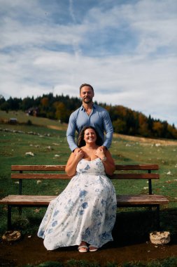 Romantik çift, manzaralı bir dağ çayırında bankta oturuyor, gülümsüyor ve birlikte huzurlu doğanın tadını çıkarıyorlar..