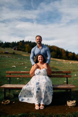 Romantik çift, manzaralı bir dağ çayırında bankta oturuyor, gülümsüyor ve birlikte huzurlu doğanın tadını çıkarıyorlar..