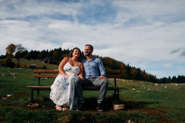 Romantik çift, manzaralı bir dağ çayırında bankta oturuyor, gülümsüyor ve birlikte huzurlu doğanın tadını çıkarıyorlar..