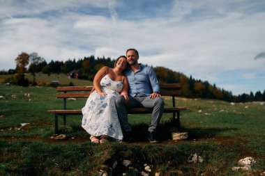 Romantik çift, manzaralı bir dağ çayırında bankta oturuyor, gülümsüyor ve birlikte huzurlu doğanın tadını çıkarıyorlar..