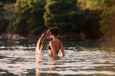 Plajda altın saat boyunca mayolu genç bir kadın ıslak saçlarını denize savuruyor. Gün batımında ıslak saçlarını suya atan bir kadın.