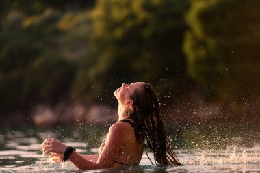 Plajda altın saat boyunca mayolu genç bir kadın ıslak saçlarını denize savuruyor. Gün batımında ıslak saçlarını suya atan bir kadın.