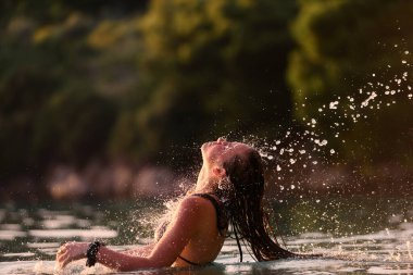 Plajda altın saat boyunca mayolu genç bir kadın ıslak saçlarını denize savuruyor. Gün batımında ıslak saçlarını suya atan bir kadın.