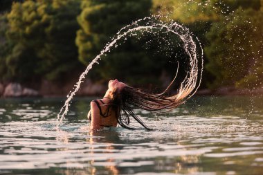 Plajda altın saat boyunca mayolu genç bir kadın ıslak saçlarını denize savuruyor. Gün batımında ıslak saçlarını suya atan bir kadın.