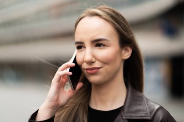 Deri ceketli genç bir kadın gülümsüyor ve elinde telefonla şehir ortamında eğleniyor..