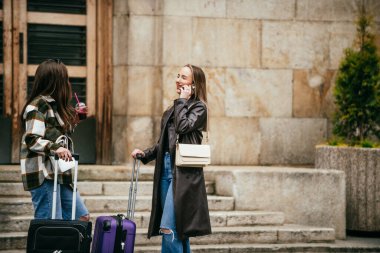 İki şık genç kadın şehir merdivenlerinde bavulları ve içkileriyle dışarıda mutlu bir şekilde konuşuyorlar..