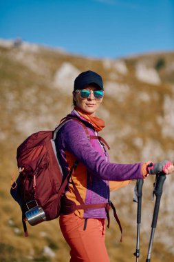 Sırt çantalı kadın yürüyüşçünün portresi. Renkli kıyafetler giyen kadın yürüyüşçünün portresi ve dağ manzarasında yürüyüş direkleri tutan güneş gözlüğü.