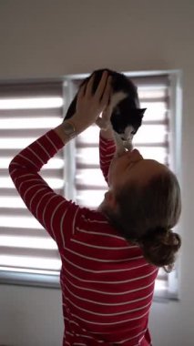 Mutlu kadın evde siyah beyaz kedi yavrusunu kaldırıyor, oyunculuğu ve evcil hayvanı sembolize ediyor.