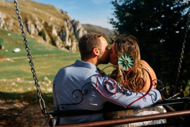Dağlarda salıncakta sallanan bir çift, arka planda büyüleyici doğal manzarayla öpüşüyorlar. Seyahat, romantizm, yaşam tarzı ve macera temaları için mükemmel