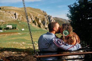Dağlarda salıncakta sallanan bir çift, arka planda büyüleyici doğal manzarayla öpüşüyorlar. Seyahat, romantizm, yaşam tarzı ve macera temaları için mükemmel
