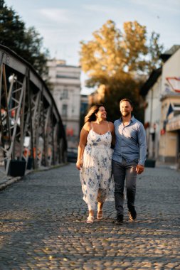 Bir kaldırım taşı caddesinde birlikte yürüyen romantik bir çift, birbirlerine sarılıyorlar ve sevgiyle gülümsüyorlar. Yaşam tarzı, romantizm, seyahat ve ilişki temaları için mükemmel