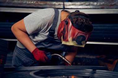 Skilled factory worker welding metal parts while wearing protective mask, gloves, and safety gear in an industrial workshop with sparks flying