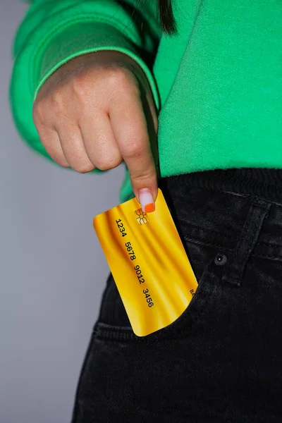 A Gold bank card is ready to be pocketed by a young lady in black pants and a green sweater on a gray background. High quality photo
