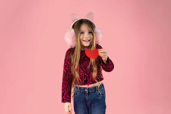 Kulaklarında pembe kulaklıklar takan, kot pantolon ve pembe baskılı süveter giyen uzun sarı saçlı küçük bir kız pembe bir arka planda küçük bir kağıt kalp tutuyor ve gülümsüyor. Yüksek kalite fotoğraf