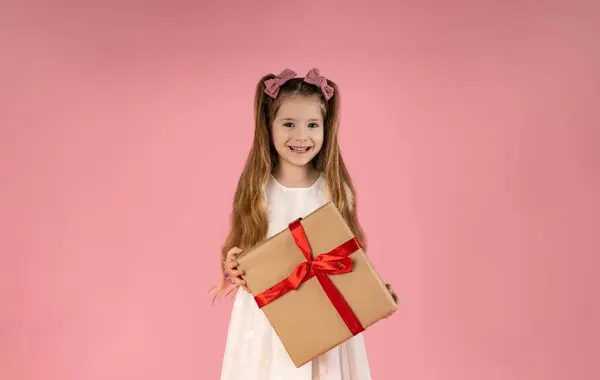 a little blonde girl with long hair holds a gift in her hands and smiles on a pink background. The girl looks out from behind the gift. The concept of a birthday party. High quality photo