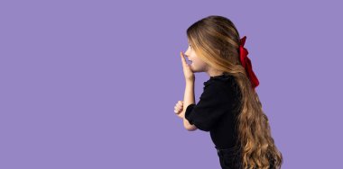 Stüdyoda mor tişörtlü, sarı saçlı, sevimli küçük bir kızın yatay görüntüsü. Mor bir duvarda poz veriyor. Şaşırmış bir ifadeyle ve ağızdan ağıza bakıyor.