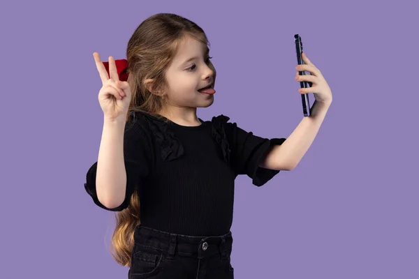 Siyah tişörtlü sevimli küçük bir kızın modern cihazla özçekim çekerken çekilmiş fotoğrafı. İzole edilmiş mor arka plan ve V işareti. Yüksek kalite fotoğraf