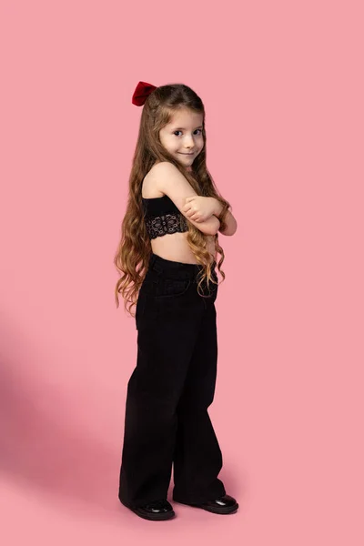 Şuna bir bak. Elleri katlanmış, siyah kot giymiş, pembe stüdyo arka planında izole edilmiş, dizayn veya sunum için boş alan gösteren gülümseyen küçük tatlı bir kız çocuğu. Yüksek