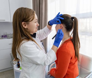 Güzel esmer bir kadının piercing ameliyatına hazırlanışının yan görüntüsü ve mavi steril eldivenli bir doktorun kalemle işaretlediği küpenin olacağı yer. Yüksek kalite fotoğraf