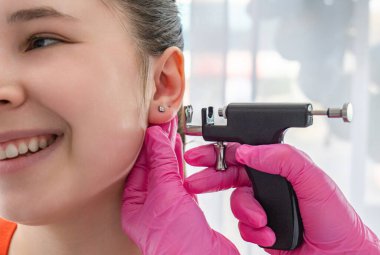 Pembe eldivenler üzerine estetik uzmanı delici silahla gülümseyen bir kızın kulağını deliyor. Yüksek kalite fotoğraf