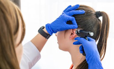 Genç bir esmerin kulak delme ameliyatı olurken yan görüntüsü. Doktorlar mavi eldivenlerle kulak delen silahı tutuyorlar. Sağlık görevlisi bir delik açar ve küpeyi kulak memesine yerleştirir.