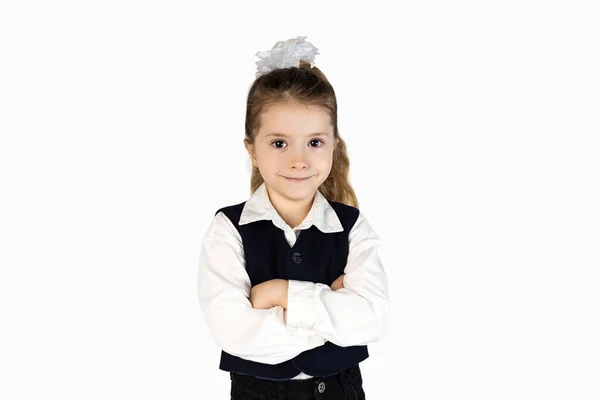 Okul üniformalı küçük bir kız, gömlek, ceket ve kravat ile tamamlanmış, kolları çapraz duran, yüzünde bir gülümseme olan.