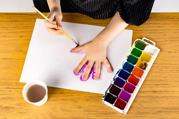 A person is painting a hand with watercolors on a piece of paper placed on a hardwood table. The paintbrush is held delicately between their fingers