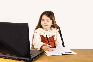 Genç bir kız, ofis malzemeleriyle çevrili bir dizüstü bilgisayar ve defterle bir masada oturuyor. Tahta panelli bir odada kişisel bilgisayarı üzerinde çalışıyor.