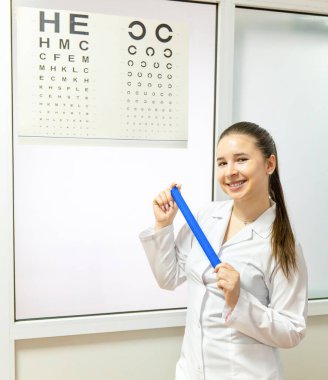 Elektrikli mavi laboratuvar önlüklü bir kadın cetvel tutarken göz çizelgesi önünde gülümsüyor. Sağlık hizmetleri odasındaki çizelgeye doğru hareket ediyor. Kapıda iş üniformasını gösteriyor.