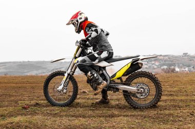 Kasklı bir motosiklet yarışçısı arazi motosikleti sürüyor. Araba lastikleri ve tekerlekleri var. Gökyüzünün altındaki toprak bir yolda.