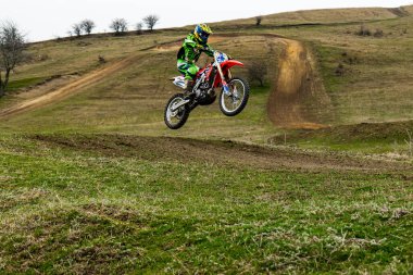 Motosikletli bir motosiklet yarışçısı havada süzülüyor, korunmak için kask takıyor. Aracın tekerlekleri ve lastikleri motorkros olayında sıçrama yaparken dönüyor.